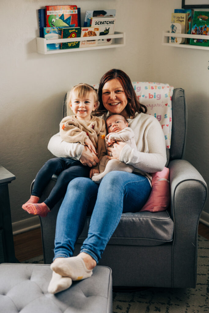 Mother smiles while holding her newborn and toddler together in a cozy nursery chair.