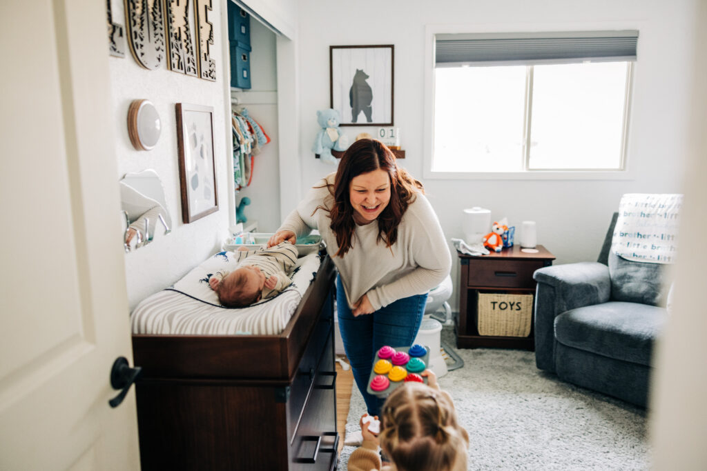 Mother smiles at her newborn on a changing table as a toddler walks over carrying toy cupcakes in a bright nursery.