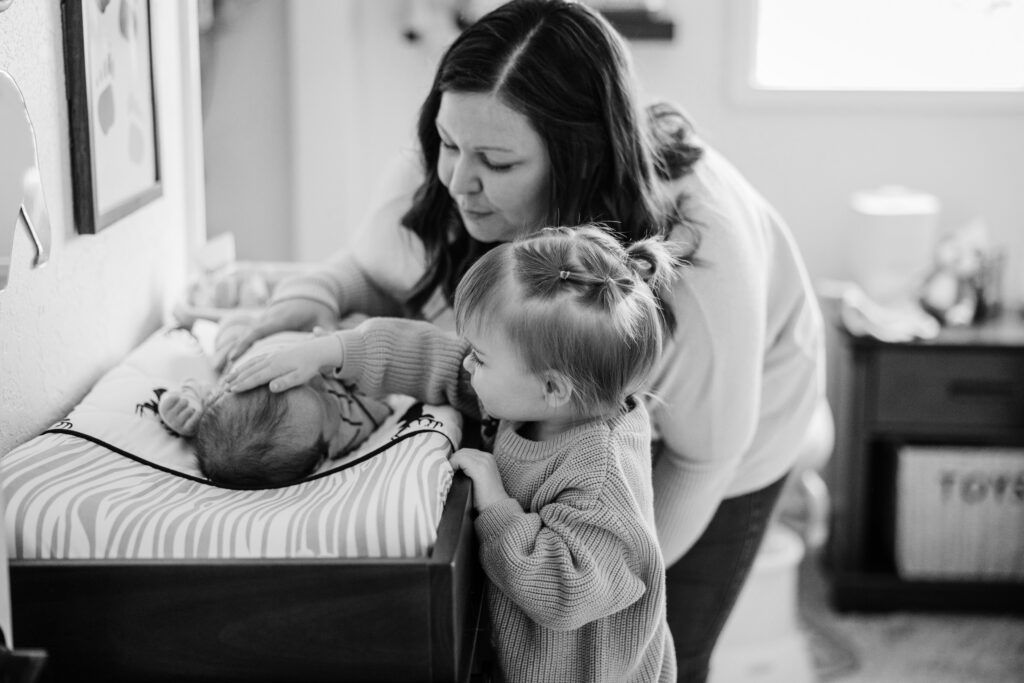 Toddler softly rests a hand on a newborn's head while mother watches beside the changing table.
