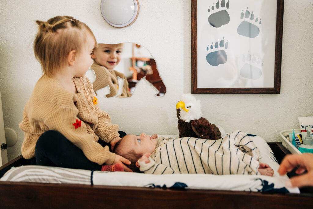 Toddler sits beside a newborn on a changing table, watching the baby near a nursery mirror.