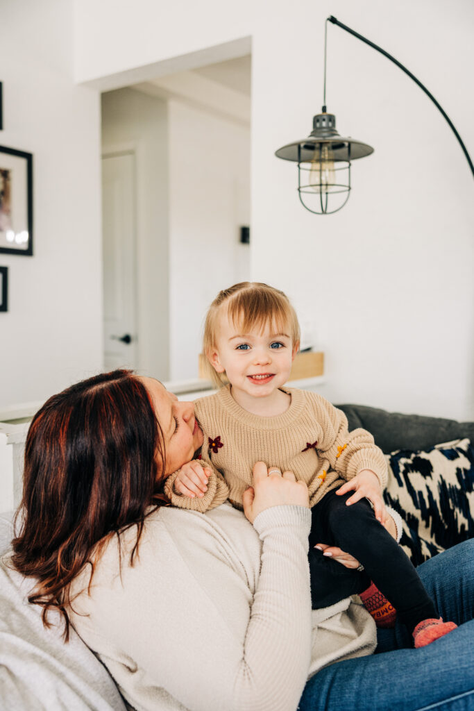 Mother holds a smiling toddler on her lap in a bright living room during a lifestyle family session.