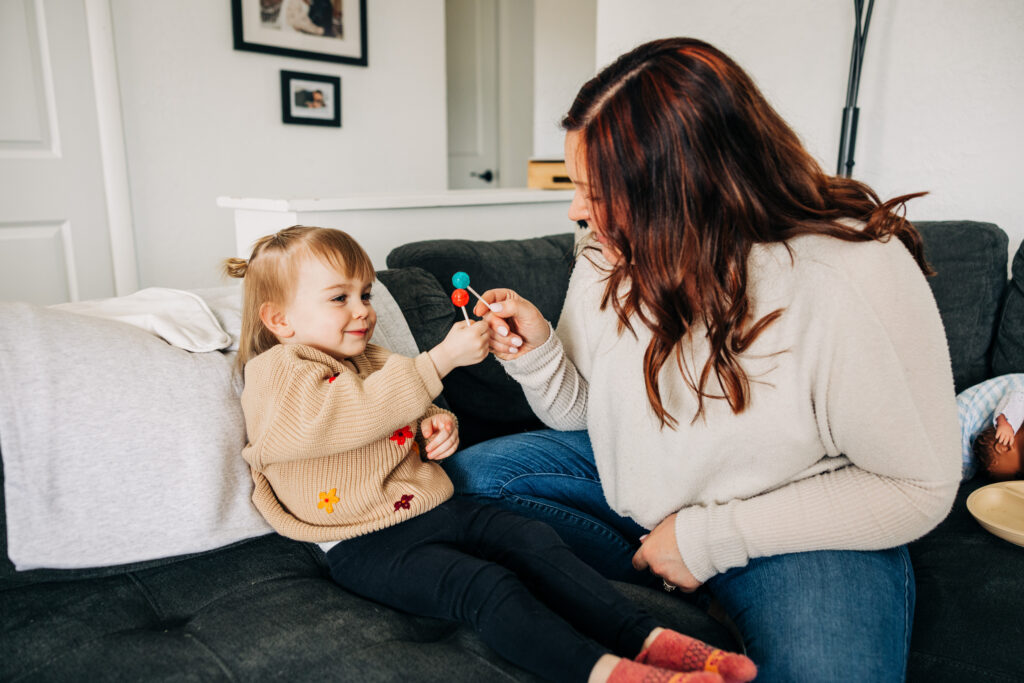 Toddler offers a colorful lollipop to mom while they sit together on the living room couch.