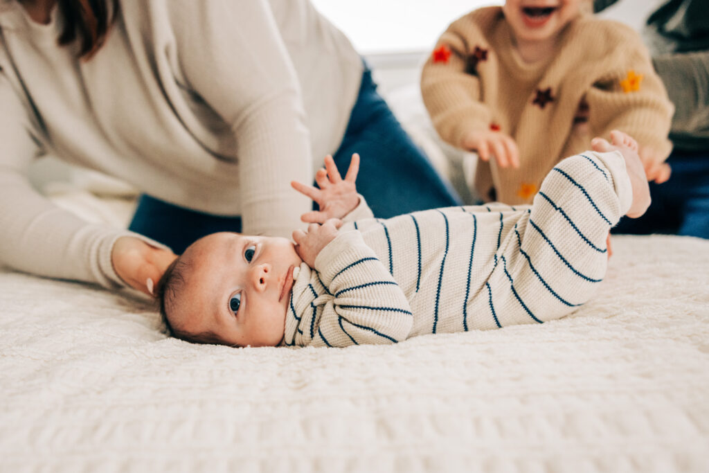 Newborn lies on a bed looking toward the camera while family members gather nearby in soft focus.