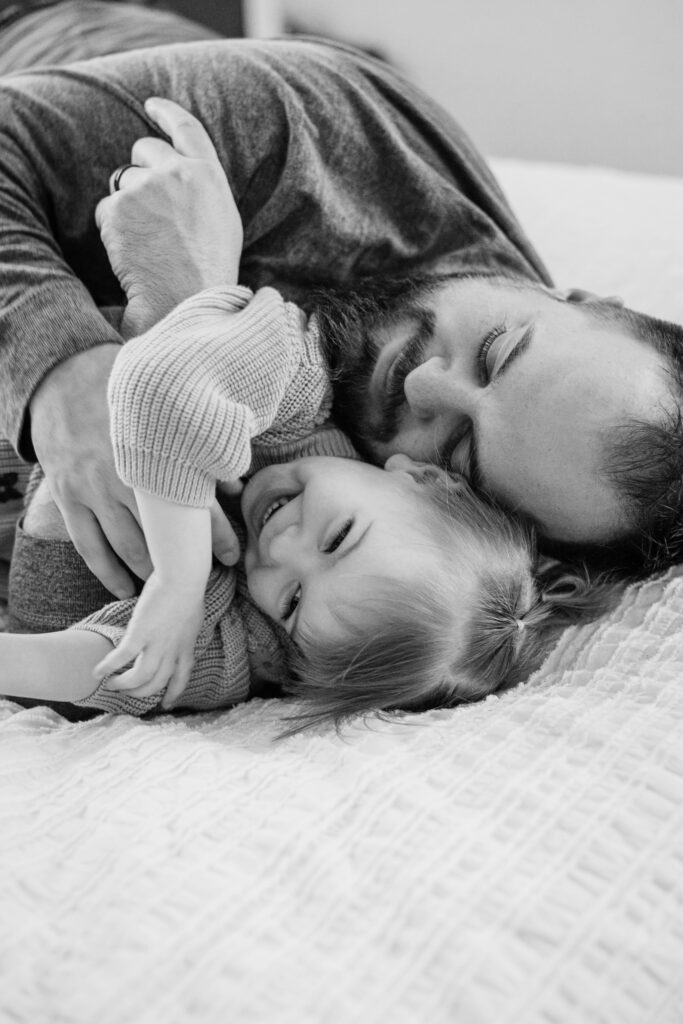 Father cuddles a smiling toddler on a bed, capturing a playful family moment in black and white.