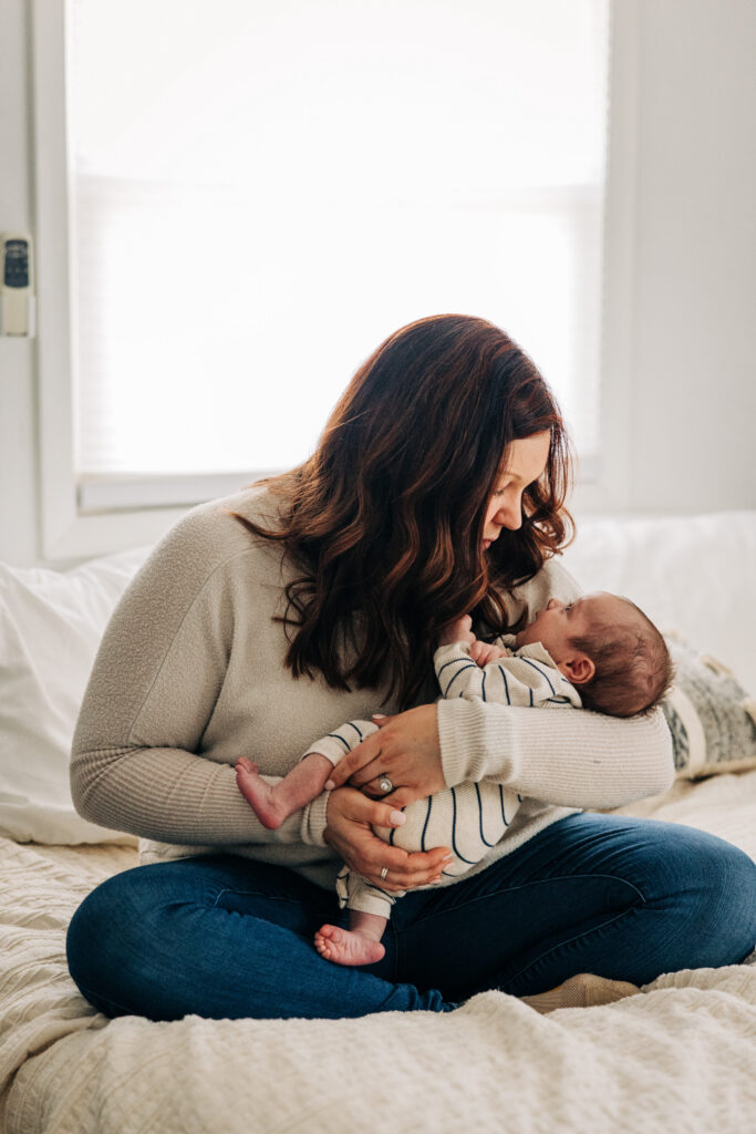 Mother sits on a bed holding her newborn, sharing a quiet moment in natural window light at home.