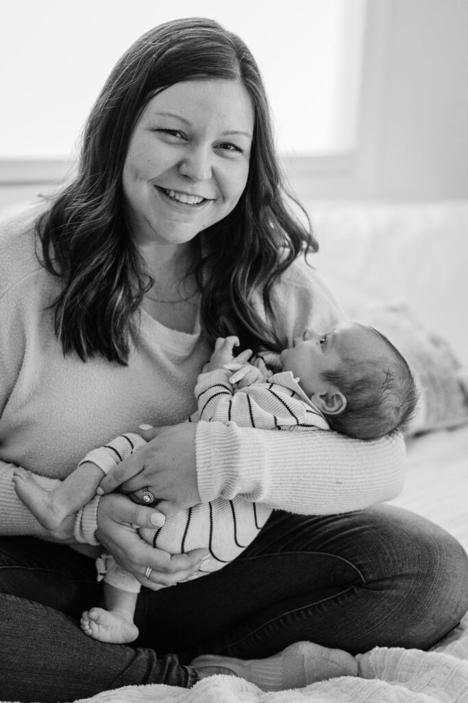 Mother smiles toward the camera while holding her newborn during a cozy in-home lifestyle newborn session.