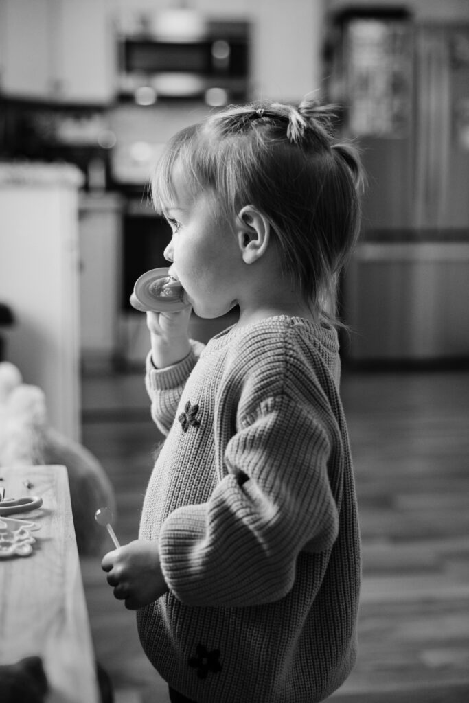 Toddler stands in a kitchen holding a lollipop, captured in a candid black-and-white lifestyle portrait.