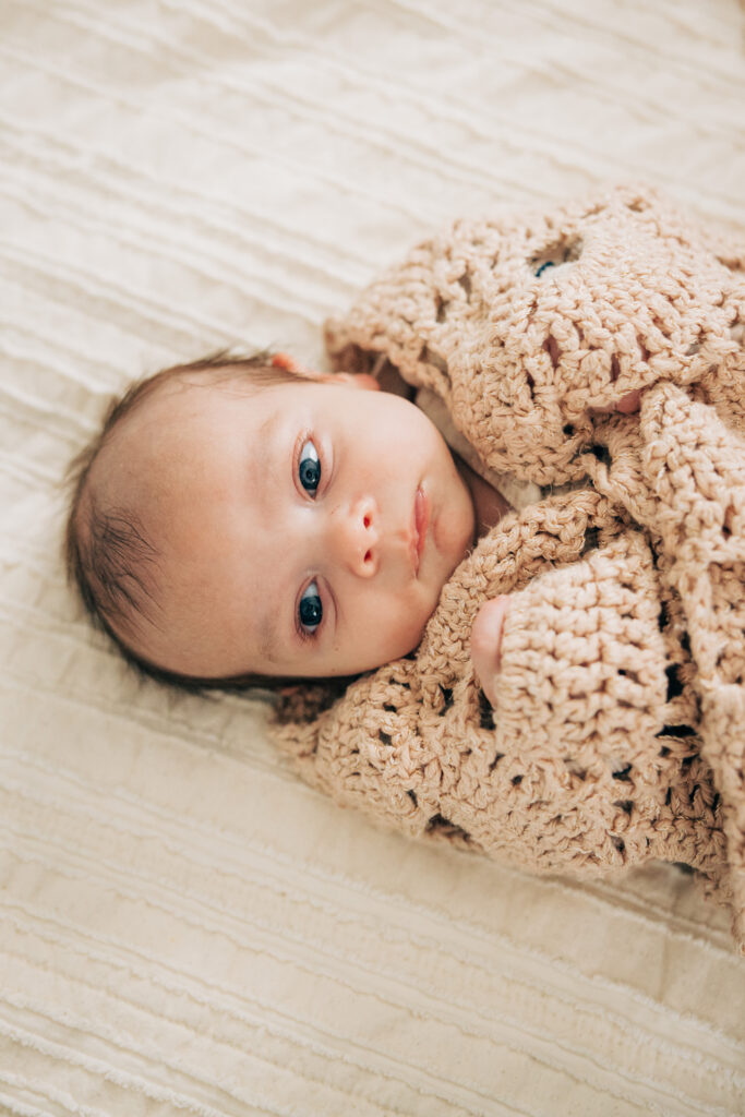Newborn lies awake on a bed wrapped in a soft knitted blanket during a cozy in-home lifestyle newborn session.