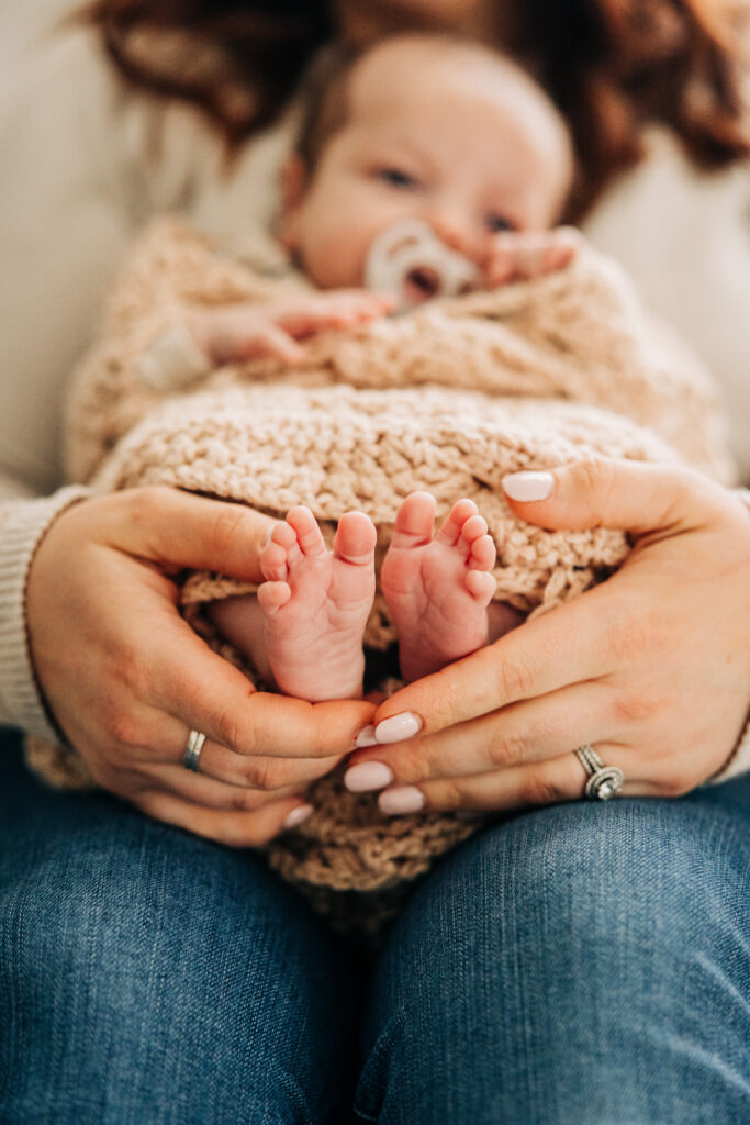 Mother cradles a swaddled newborn, highlighting the baby's tiny feet during a natural in-home lifestyle newborn session.