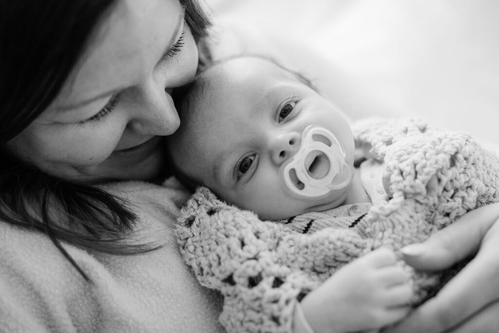 Mother cuddles her newborn wrapped in a knitted blanket as the baby looks toward the camera with a pacifier during an in-home lifestyle newborn session.
