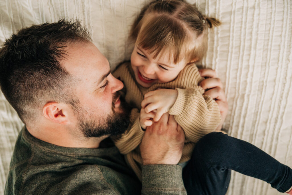Man holding a laughing toddler while they play together on a bed.