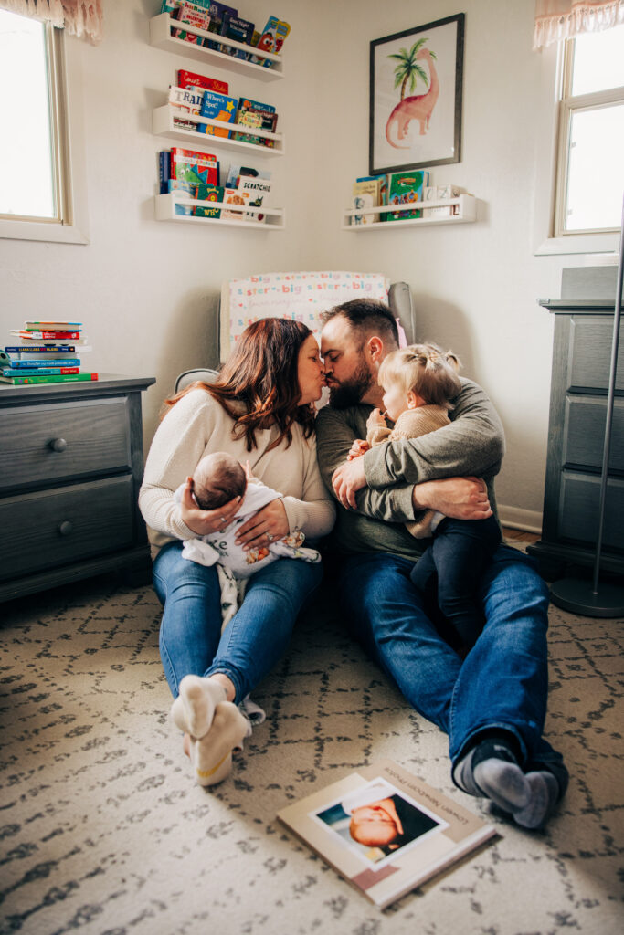 Parents sitting on a nursery floor holding their newborn and toddler while sharing a kiss.